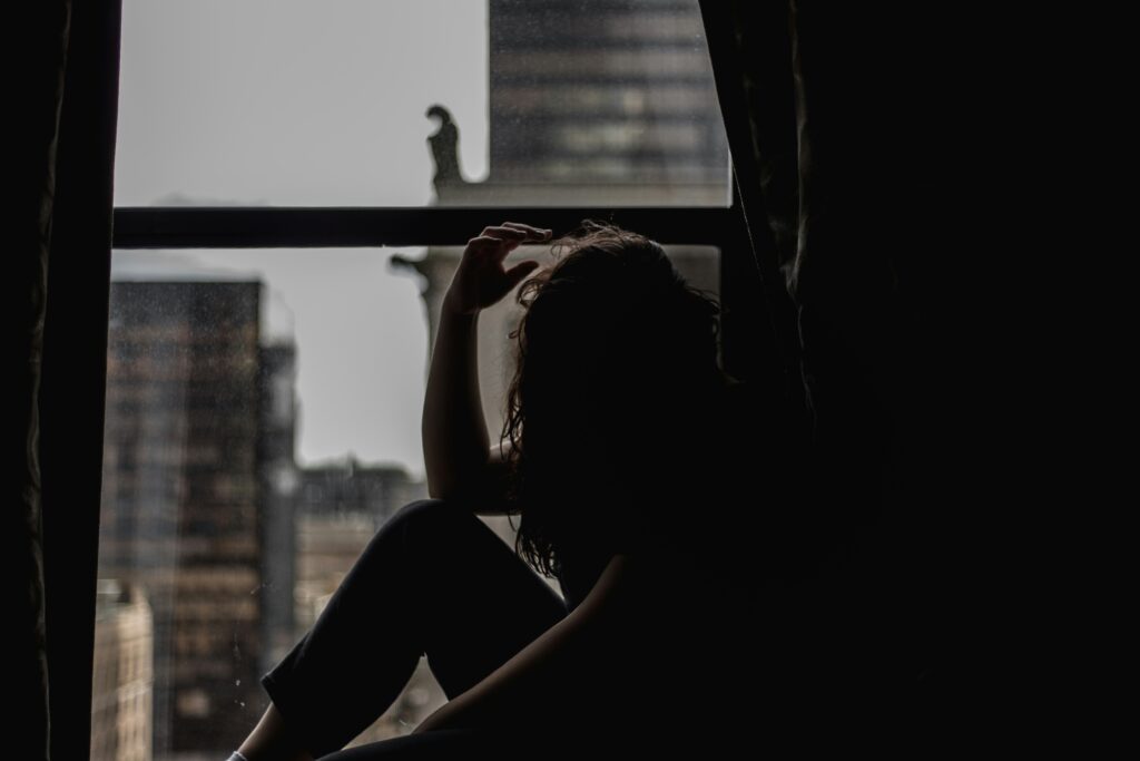Femme assise dans l'ombre près d'une fenêtre, symbole de solitude, de douleur intérieure et de lutte silencieuse.