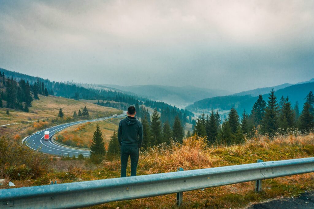 A man stands beside a winding road and looks out over the mountainous landscape.